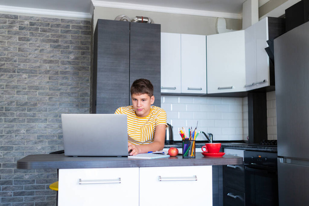Cute młody nastolatek w białej koszuli siedzi za biurkiem w kuchni obok laptopa i badania. Poważny chłopak w słuchawkach odrabia lekcje słuchania. Dom, edukacja na odległość, samodzielne uczenie się przez dzieci. - Zdjęcie, obraz