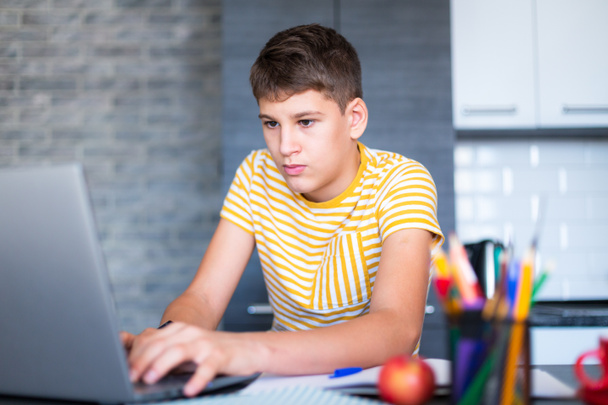 Χαριτωμένος νεαρός έφηβος με λευκό πουκάμισο κάθεται πίσω από το γραφείο στην κουζίνα δίπλα στο laptop και μελέτη. Σοβαρό αγόρι με ακουστικά κάνει εργασίες, μάθημα ακρόασης. Σπίτι, εξ αποστάσεως εκπαίδευση, αυτο μελέτη από τα παιδιά. - Φωτογραφία, εικόνα