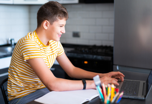 Cute young teenager in white shirt sitting behind desk in kitchen next to laptop and study. Serious boy in earphones makes homework, listening lesson. Home, distance education, self study by kids. - Foto, Imagem