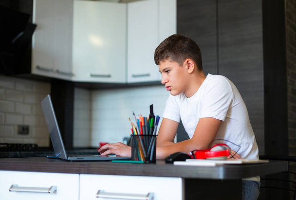 Cute młody nastolatek w białej koszuli siedzi za biurkiem w kuchni obok laptopa i badania. Poważny chłopak odrabia lekcje słuchania. Dom, edukacja na odległość, samodzielne uczenie się przez dzieci. - Zdjęcie, obraz