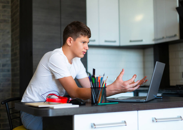 Roztomilý mladý teenager v bílé košili sedí za stolem v kuchyni vedle notebooku a studovat. Vážný chlapec dělá úkoly, poslouchá lekce. Home, distanční vzdělávání, samostudium dětmi. - Fotografie, Obrázek