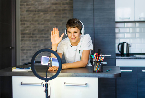 Nette fröhliche Junge Blogger Aufnahme auf Smartphone-Video auf seiner Küche für Website. Teenager in weißem Hemd und Kopfhörer, die zu Hause Videos drehen. Social Media-Konzept. - Foto, Bild