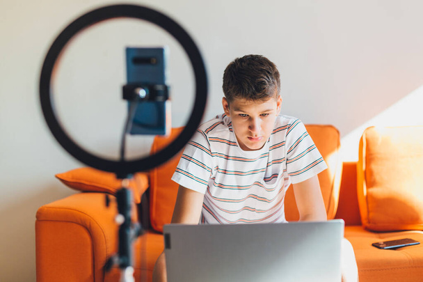 Νεαρός έφηβος blogger ηχογραφεί βίντεο στο σπίτι. Χαρούμενο χαριτωμένο αγόρι που δείχνει με τα χέρια σε ένα smartphone. Αγόρι με κάμερα να καταγράφει βίντεο στον πορτοκαλί καναπέ. Έννοια μέσων κοινωνικής δικτύωσης. - Φωτογραφία, εικόνα