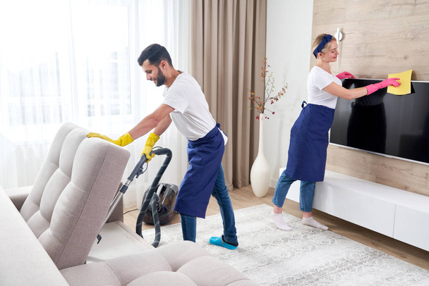 Mavi üniformalı profesyonel temizlikçiler dairenin oturma odasındaki mobilyaların tozunu siliyorlar. Temizlik hizmeti kavramı - Fotoğraf, Görsel