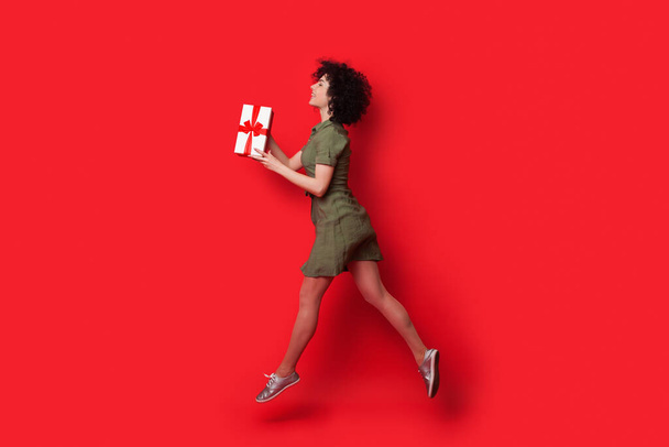Mujer caucásica delgada con el pelo rizado corriendo en una pared de estudio rojo con un regalo dándoselo a alguien - Foto, imagen