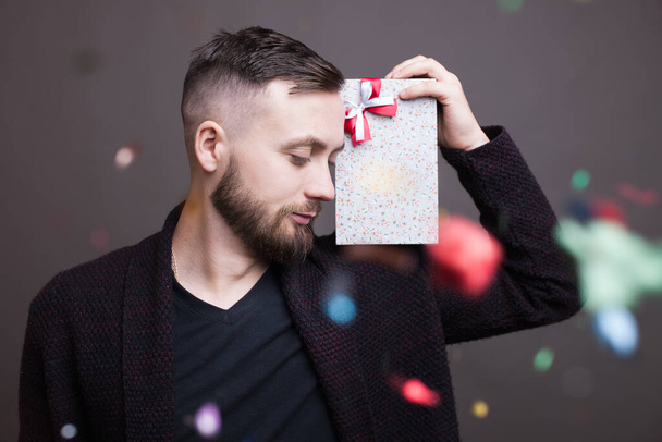 Bearded caucasian man with a present on his shoulder is posing in formal wear on a gray studio wall with confetti - 写真・画像