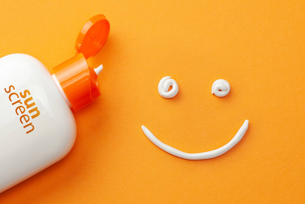Sunscreen on orange background. Plastic bottle of sun protection and white cream in the shape of Smiley, smiling face - Photo, Image