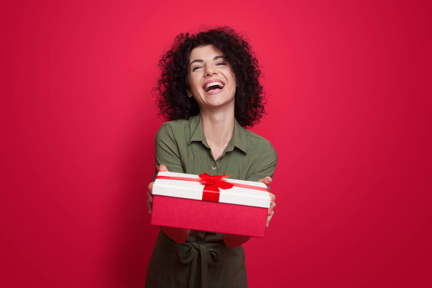 Charming brunette woman with curly hair giving a present at camera smiling on a red studio wall - Zdjęcie, obraz