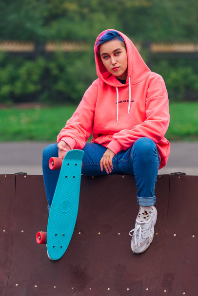 Portrait of a trendy pretty young girl dressed in pink hoodie and jeans sitting next to the skateboard coart with her blue plastic skateboard. - Фото, зображення