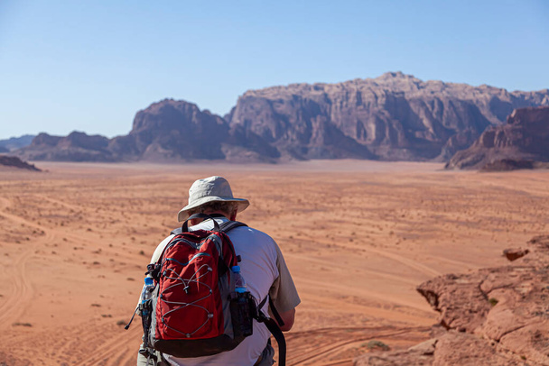Un turista de mediana edad occidental con una mochila roja y sombrero solar está de pie en el borde de un acantilado de piedra arenisca mirando hacia abajo para te vasto paisaje del desierto en el pintoresco Wadi Rum, Jordania. - Foto, imagen