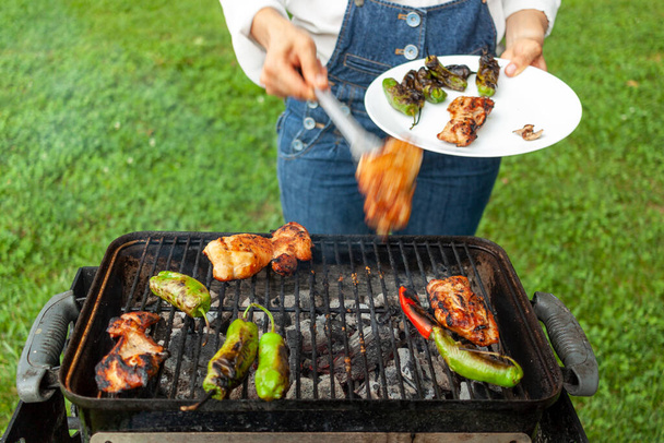 a chef wearing jean overalls and white shirt is by a charcoal grill where marinated chicken slices, red and green peppers are fire grilled. She uses tongs to flip them and collect them into a plate. - Photo, Image