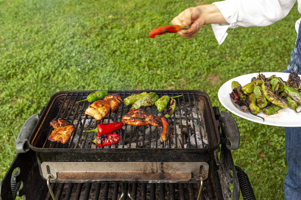 Ein Koch in Jeanshose und weißem Hemd steht vor einem Holzkohlegrill, auf dem marinierte Hühnerscheiben, rote und grüne Paprika gegrillt werden. Sie verwendet eine Zange, um sie zu drehen und in einem Teller zu sammeln. - Foto, Bild