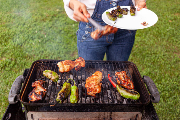 a chef wearing jean overalls and white shirt is by a charcoal grill where marinated chicken slices, red and green peppers are fire grilled. She uses tongs to flip them and collect them into a plate. - Photo, Image