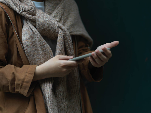 Вид сбоку женщины в свитере, использующей смартфон, стоя на темном фоне  - Фото, изображение