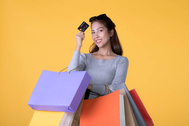 Beautiful young Asian woman with colourful shopping bags and credit card isolated on yellow background - Φωτογραφία, εικόνα