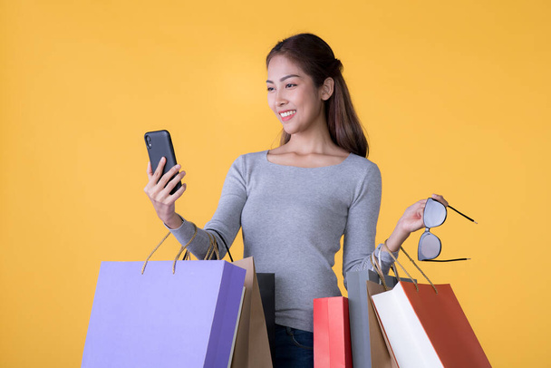 Beautiful young Asian woman with colourful shopping bags and smartphone isolated on yellow backgroun - Fotografie, Obrázek