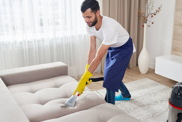 Close-up of housekeeper holding modern washing vacuum cleaner and cleaning dirty sofa with professionally detergent. Professional springclean at home concept - Fotó, kép