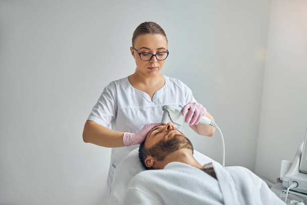 Naiskosmetologi hoitaa miehen ihoa laserlaitteella - Valokuva, kuva