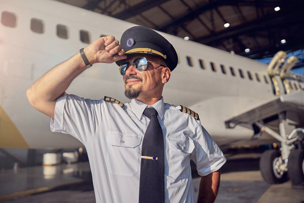 Magabiztos szakállas férfi tartja pilóták kalap a fejét, miközben küld időt a szabadban - Fotó, kép