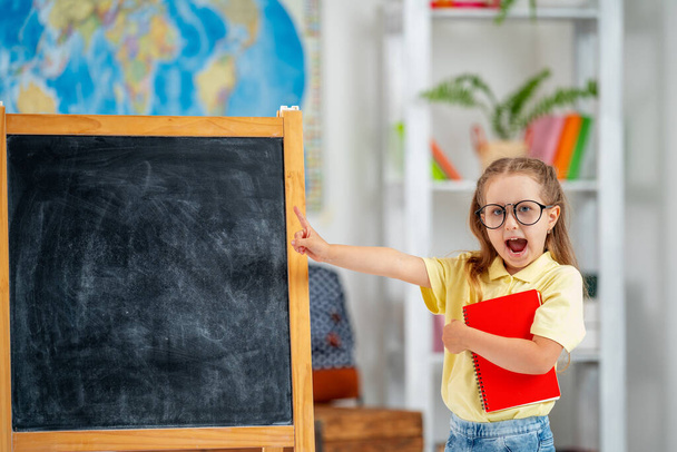Back to school. funny smiling schoolgirl in glasses with book in her hand, pointing at blackboard. child poses in school classroom against blackboard. Preparation for school. Education. Copy space. - Photo, Image