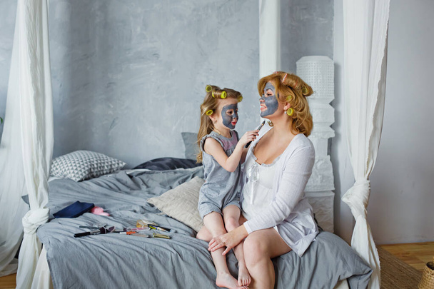 母親と子供は一緒に化粧品の手順を行います。幸せな家族の母親と小さな娘がベッドの上に座ってベッドルームで顔マスクを作ります.エステの概念,スパとウェルネスセンター. - 写真・画像