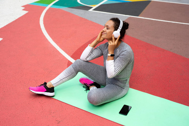 De jonge vrouw in een grijs trainingspak zit op een groene mat met een koptelefoon en geniet van de muziek - Foto, afbeelding