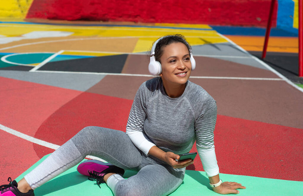 The young woman in gray tracksuit sits on green mat in headphones with smartphone in hand - Foto, immagini