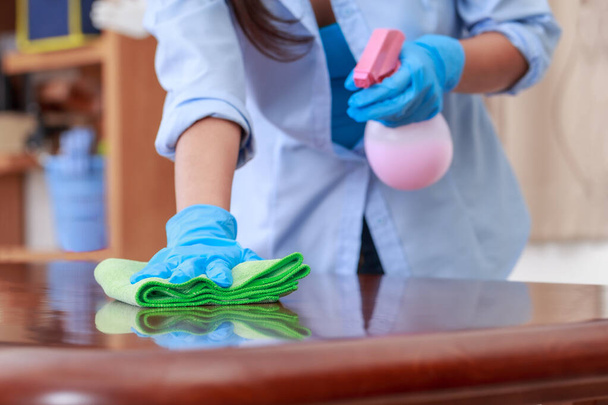 Donna con guanti protettivi blu con un panno verde per la pulizia in microfibra Pulizia di tavoli in legno e bottiglie spray con soluzioni igienizzanti, detergenti e disinfettanti per una buona igiene. - Foto, immagini