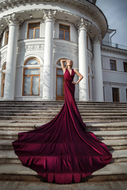 Schöne schlanke Mädchen in einem dunkelroten seidenfließenden Kleid mit einer langen Schleppe auf den Stufen des Palastes. - Foto, Bild