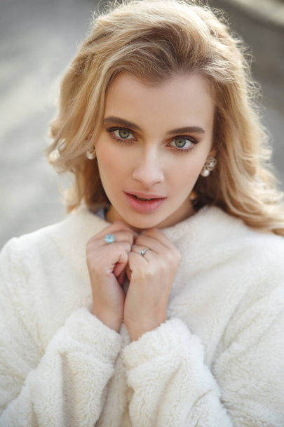 Portrait of a cute blonde girl in a cozy white fur coat outside. - Фото, изображение