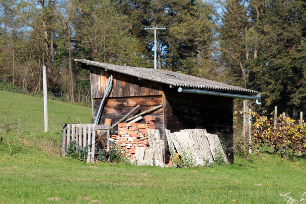 Старые деревянные доски сарай инструмент, покрытый ветхой черепицы крыши в окружении строительного материала и травы на стороне небольшого холма с высокими деревьями на заднем плане - Фото, изображение