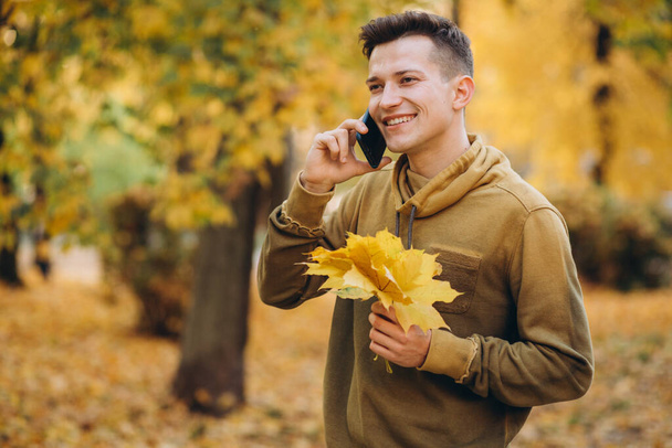 Портрет красивого і щасливого хлопця, який посміхається і розмовляє по телефону в осінньому парку
 - Фото, зображення