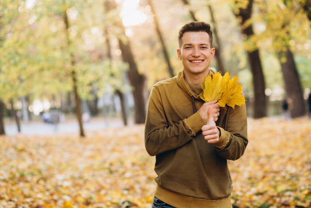 Retrato de chico guapo sonriendo y sosteniendo un ramo de hojas de otoño en el parque - Foto, Imagen