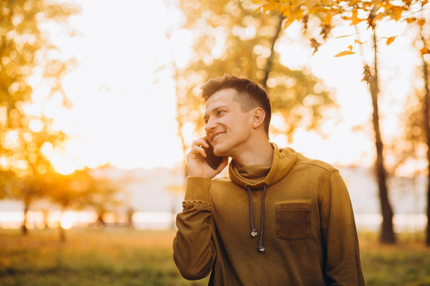Portrait of handsome and happy guy smiling and talking on the phone in the autumn park - Photo, Image