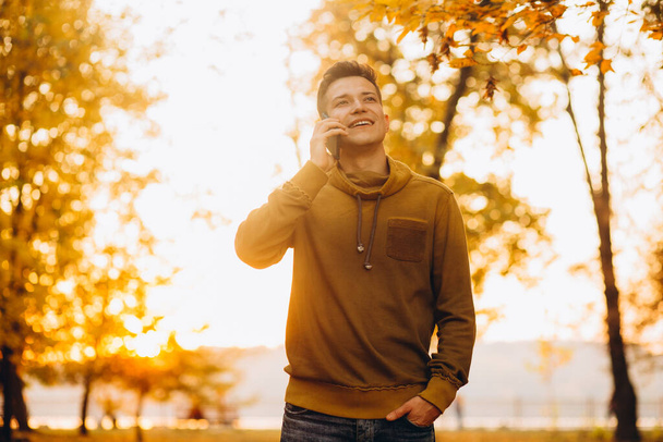 Портрет красивого и счастливого парня, улыбающегося и разговаривающего по телефону в осеннем парке - Фото, изображение
