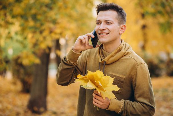 Портрет красивого і щасливого хлопця, який посміхається і розмовляє по телефону в осінньому парку
 - Фото, зображення