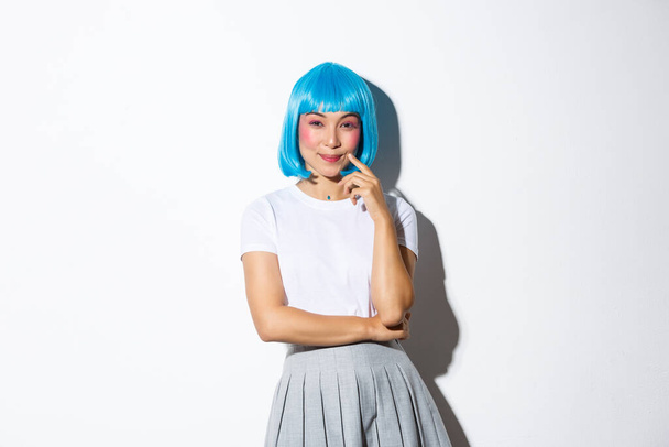 Image of cunning smiling asian girl in blue party wig, looking devious at camera, have an idea, standing over white background - Photo, Image