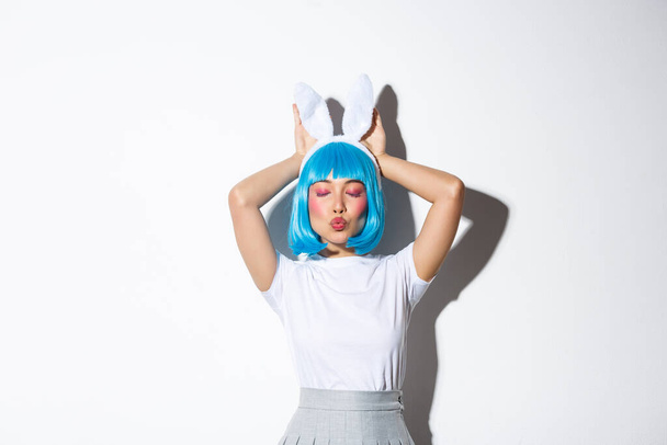 Linda menina asiática em azul peruca curta e orelhas de coelho, bebendo coquete e posando sobre fundo branco no traje de halloween - Foto, Imagem