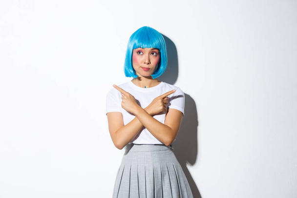 Indecisive cute asian girl making choice, pointing fingers sideways, wearing halloween costume and blue short wig, standing over white background - Photo, Image