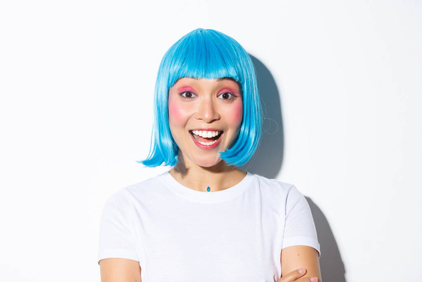 Imagen de la excitada chica asiática linda en traje de halloween y peluca azul, sonriendo divertido, de pie sobre fondo blanco - Foto, Imagen