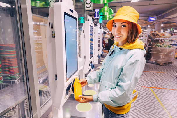 Szczęśliwa młoda klientka kupuje i płaci za butelkę soku pomarańczowego przy kasie automatu samoobsługowego w nowoczesnym supermarkecie, aby uniknąć czekania w kolejce - Zdjęcie, obraz