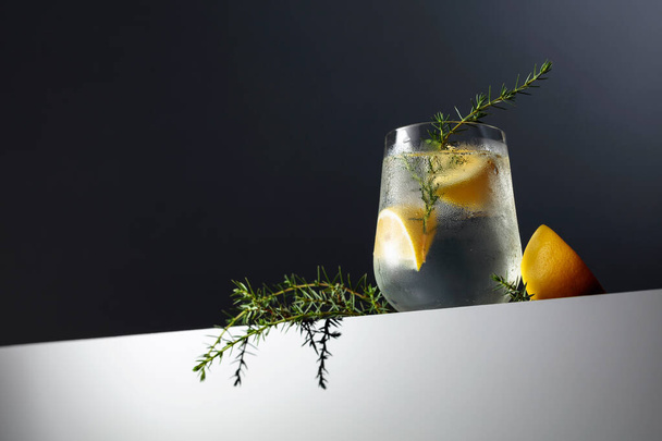 Alcoholdrank (gin tonic cocktail) met citroen, jeneverbes tak, en ijs op een donkere reflecterende achtergrond, kopieer ruimte. Iced cocktail drankje met citroen.  - Foto, afbeelding
