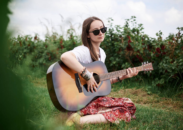 Young brunette hippie woman, wearing boho style clothes, sitting on green grass, holding guitar. Indie musician relaxing on green currant field on sunny summer day. Eco tourism concept. - Photo, image