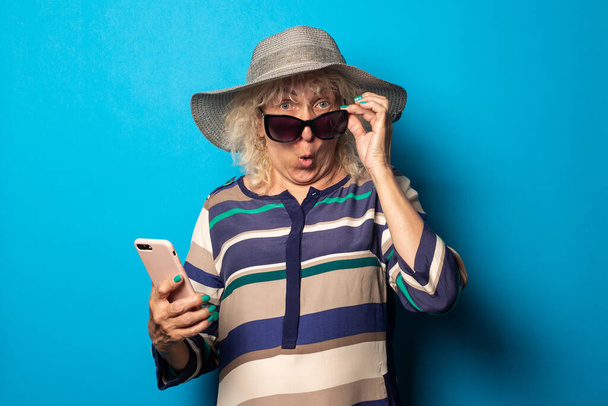Gözlüklü ve şapkalı yaşlı kadının mavi arka planda telefonu tutmasına şaşırdım. - Fotoğraf, Görsel