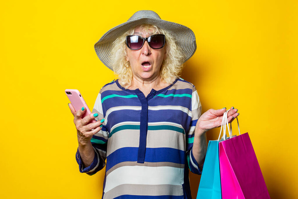 Şapkalı ve gözlüklü, şaşırmış yüzlü yaşlı kadın, sarı arka planda alışveriş torbaları ve telefon tutuyor.. - Fotoğraf, Görsel