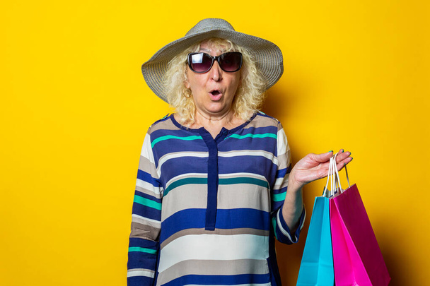 Old woman with surprised face in hat and glasses holds shopping bags with purchases on yellow background. - Fotó, kép