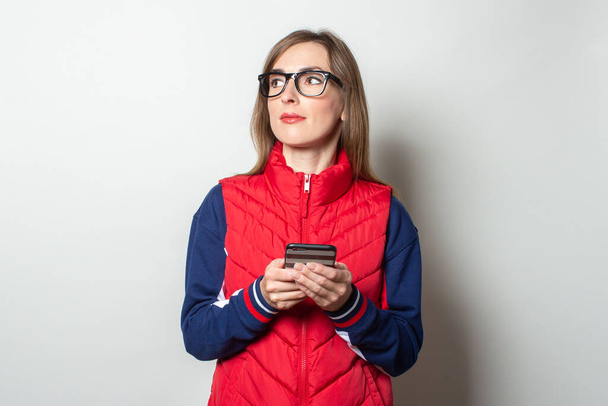 Young woman looks to the side in a red vest holding her phone in her hands on a light background. - Foto, afbeelding