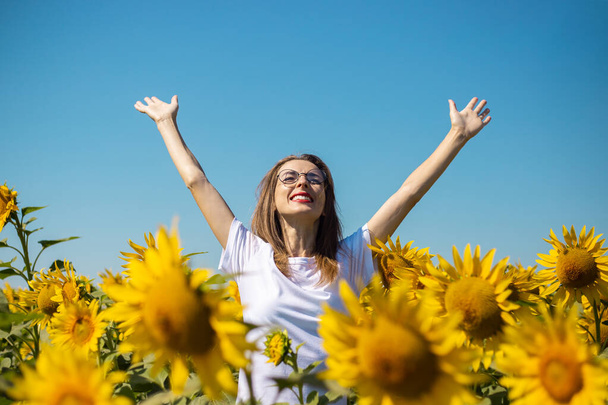 Young woman in a white t-shirt and glasses with raised up hands on a sunflower field on a summer sunny day. - Photo, Image