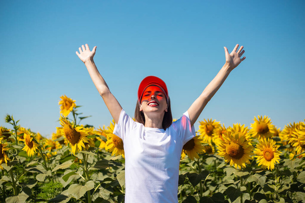 Mujer joven con una visera de sol roja y una camiseta blanca extiende sus manos hacia el cielo y se encuentra con el amanecer en un campo de girasol. - Foto, Imagen