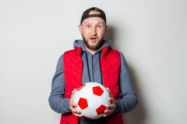 Νεαρός άνδρας με ένα έκπληκτο πρόσωπο σε ένα καπέλο, κουκούλα και γιλέκο κρατώντας μια μπάλα ποδοσφαίρου σε ελαφρύ φόντο. - Φωτογραφία, εικόνα